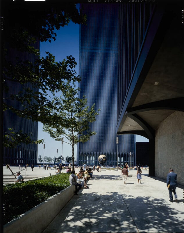 A sunny outdoor plaza featuring the original World Trade Center twin towers. People are walking or sitting on benches. A globe sculpture is visible between the two towers. The sky is clear, and a large tree provides shade.