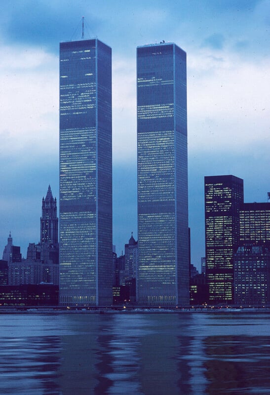 The image shows the Twin Towers of the World Trade Center in New York City. The iconic skyscrapers dominate the skyline, with surrounding buildings visible, all illuminated against a dusk sky with their lights reflecting in the water below.