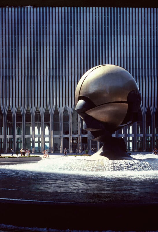 A large bronze sculpture called "The Sphere" stands at the center of a fountain with water cascading around it. In the background, there's a tall building with a unique facade featuring curved vertical lines and arch-like windows. People are seen walking nearby.