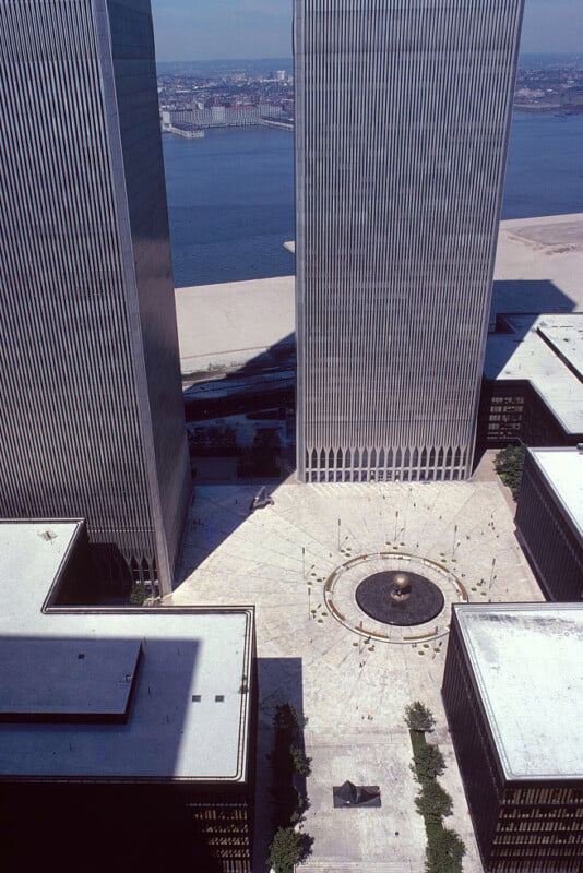 Aerial view of the World Trade Center Plaza in New York City, showing the twin towers and the circular fountain in the center. The buildings' reflections and shadows are visible on the plaza, with the Hudson River and New Jersey visible in the background.