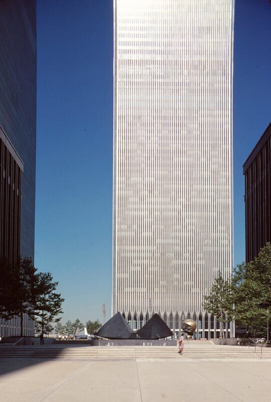 A towering glass and steel skyscraper dominates the background, part of the World Trade Center. The foreground features a large, open plaza with various sculptures, including a black geometric structure and a bronze globe. Sparse trees frame the plaza.