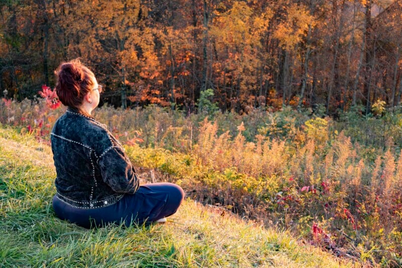 A person with sunglasses and a patterned jacket sits cross-legged on a grassy hill, overlooking a forest with trees in autumn colors. The scene is bathed in warm, golden sunlight.