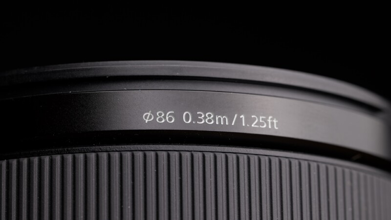 Close-up of a camera lens with etched specifications: ϕ86 0.38m/1.25ft. The lens features a textured focus ring, and the lighting highlights the details against a dark background.