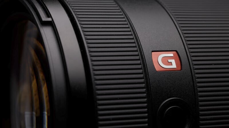 Close-up of a black camera lens with a textured grip, featuring a red and white "G" logo. The lighting highlights the lens's ridged surface, creating a dramatic and professional appearance.