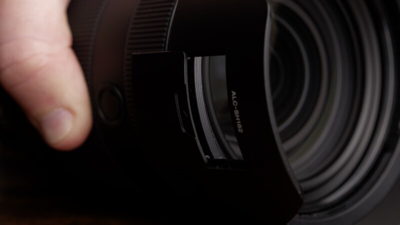 Close-up of a person holding a large camera lens with a lens hood attached. The focus is on the intricate details of the lens elements visible through the glass, emphasizing the precision and craftsmanship of the camera equipment.