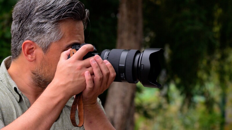 A person with gray hair holds a professional camera up to their eye, focusing on capturing an image. The background is blurred greenery, suggesting an outdoor location. The individual wears a casual, light-colored shirt.