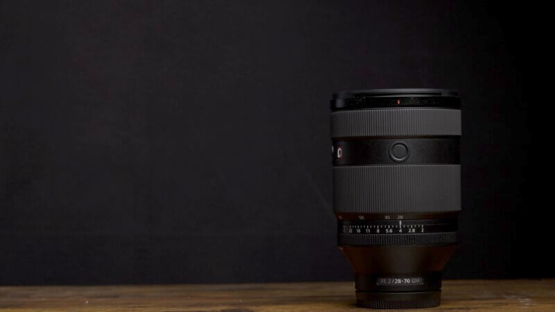 A camera lens with a smooth, dark gray body and a red logo is positioned upright on a wooden surface. The background is a solid black, creating contrast and highlighting the lens. The lens model "FE 24-70 GM" is visible near the base.