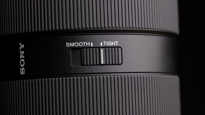 Close-up of a camera lens showing a switch labeled "Smooth" and "Tight." The switch is positioned in the middle. The lens is black with textured grip sections, and part of the brand name is visible on the side.