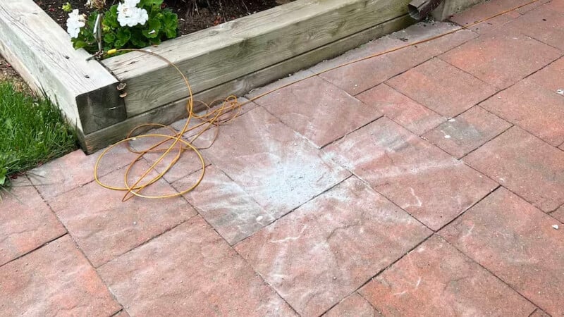 A brick patio with a circular white stain resembling a sunburst pattern. A coil of yellow extension cord is resting next to the stain. A wooden planter with white flowers is in the background.