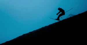 Silhouette of a person skiing downhill on a steep slope against a deep blue sky. The skier is airborne, capturing a dynamic motion.