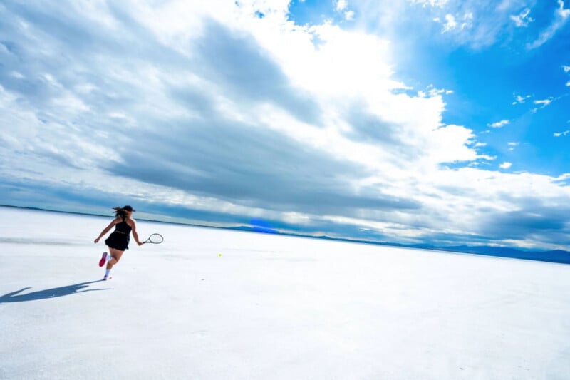 A person in athletic attire runs with a tennis racket across a vast, white, open landscape under a bright blue sky with scattered clouds. The wide expanse suggests a salt flat, with distant mountains on the horizon.