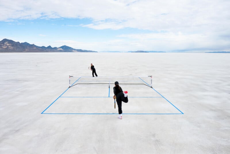 Two people play tennis on a minimalistic court marked with blue lines on a vast, flat salt plain. The sky is partly cloudy with distant mountains in the background, creating a surreal landscape.