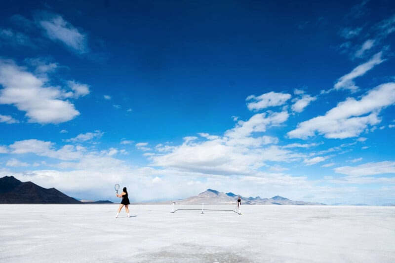 Two people play tennis on a vast, flat white surface under a bright blue sky with scattered clouds. Mountains are visible in the distance, enhancing the surreal setting.