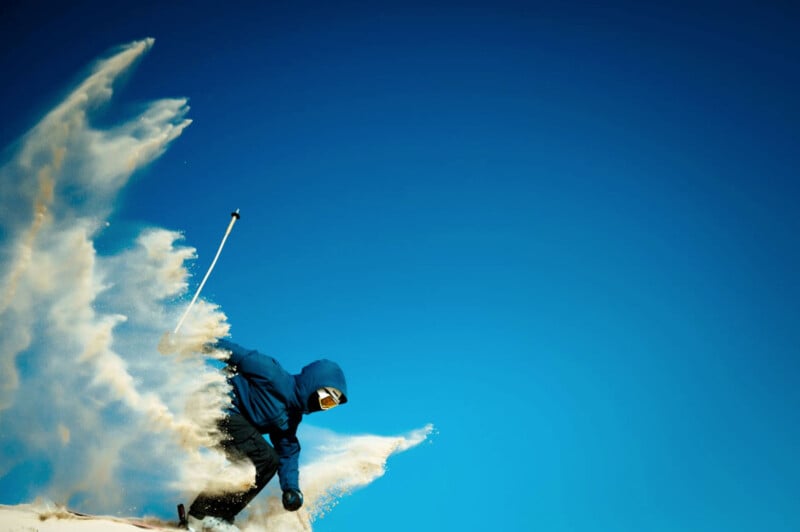 A skier in a blue outfit and goggles makes a sharp turn on a snowy slope, creating a spray of snow against a clear blue sky.