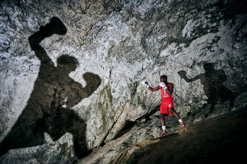 A boxer in red gear poses inside a rocky cave, casting a large, dramatic shadow on the wall. The scene is dimly lit, creating a striking silhouette effect against the textured stone backdrop.