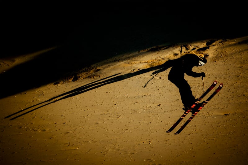 A skier in mid-air against a dark, sandy background, casting a long shadow. The image captures the dynamic motion and contrast between the illuminated sand and the silhouette of the skier.