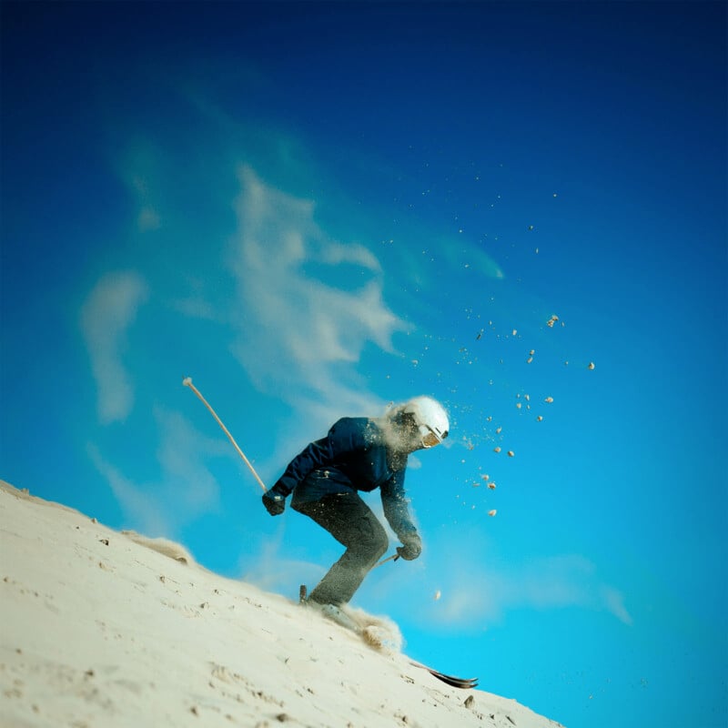 A skier wearing a helmet and goggles glides down a sand dune under a clear blue sky. Sand sprays up behind them, creating a dynamic and adventurous scene.