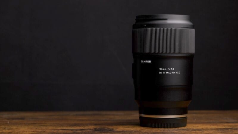 A black camera lens, labeled "Tamron 90mm F/2.8 Di III Macro VC," sits upright on a wooden surface against a dark background.