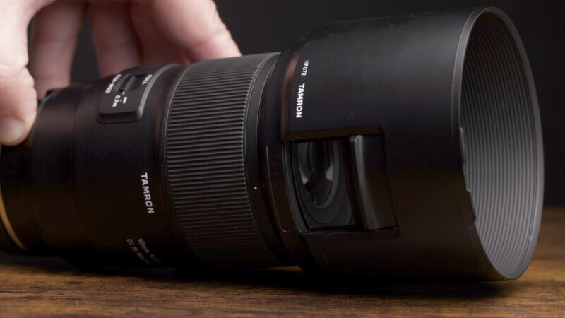 A close-up of a Tamron camera lens placed on a wooden surface is shown. A hand is gently holding the lens from the side. The lens features detailed texture on the focus ring and branding on the side.