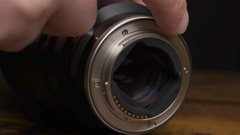 Close-up of a person adjusting a camera lens. The focus is on the lens mount, showing the metal contacts and the aperture blades inside. The background is dark, emphasizing the lens details.