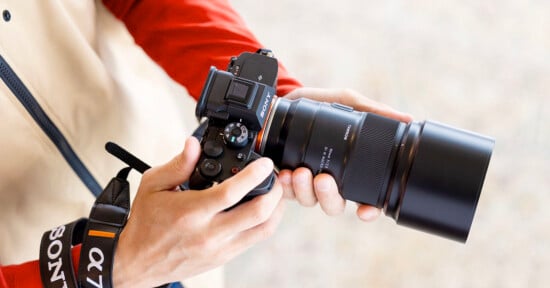 A person holding a Sony camera with a large lens, adjusting settings while wearing a camera strap labeled "Sony α7.