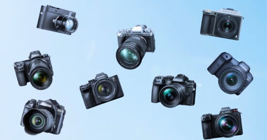 A variety of digital cameras are arranged against a blue background. The cameras include models from brands like Fujifilm, Sony, Pentax, Panasonic, and another unidentifiable brand. They are positioned in a circular pattern, showcasing their lenses prominently.