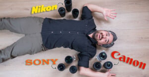 A man lies on a wooden floor surrounded by various camera lenses. The words "Nikon," "Sony," and "Canon" are written near groups of lenses. He looks up with a playful expression, wearing a black shirt and khaki pants.