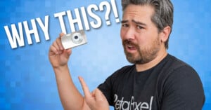 A person holds and points to a small digital camera against a blue pixelated background. Large text reads "WHY THIS?!". The person appears curious or surprised.