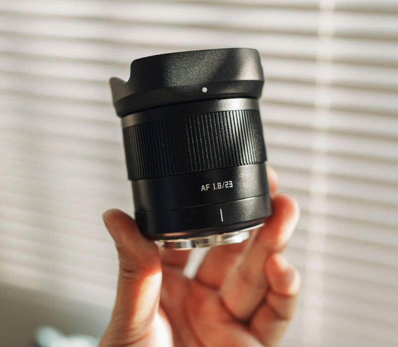 A hand is holding a black camera lens labeled "AF 1.8/23" against a background of window blinds with sunlight filtering through. The lens has a wide, textured grip and a silver metal mount.