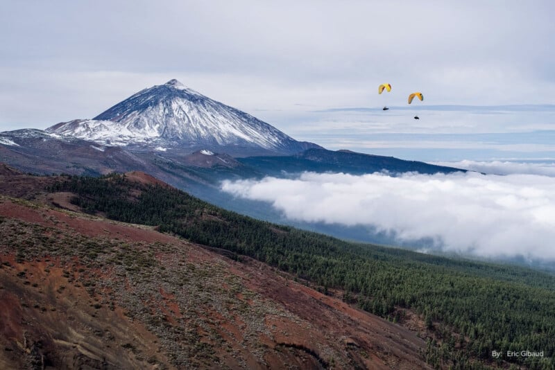 Paragliders soar above green and red volcanic landscapes, with a snow-capped mountain in the background and a layer of clouds below. The sky is overcast, adding to the dramatic scenery.