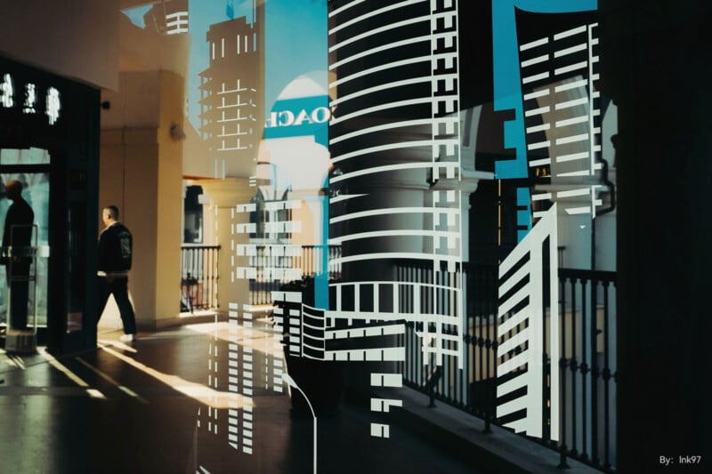 A man walks along a sunlit corridor with reflective glass. Architectural patterns overlay the scene, creating an illusion of tall buildings. Shadows and light combine, merging interior and exterior elements with urban artistry.