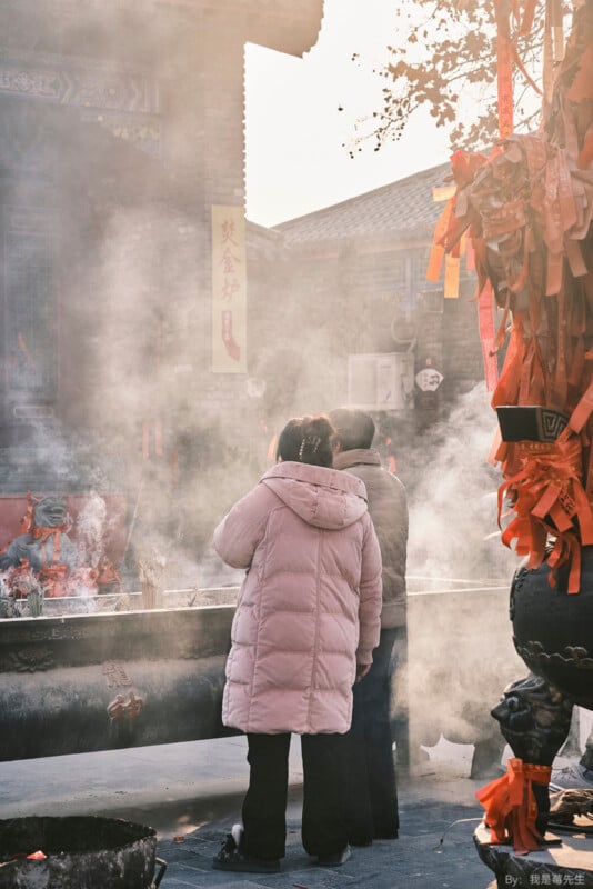 Two people stand in front of an outdoor altar, surrounded by smoke from burning incense. They appear to be engaged in a ritual or prayer. Traditional architecture and red decorations are visible in the background.