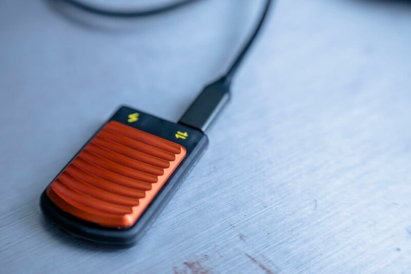 Close-up of an electronic device resembling a card reader with a ribbed orange surface, connected by a black cable. Two symbols are visible on the device: a lightning bolt and Wi-Fi signal. The background is a smooth, metallic surface.