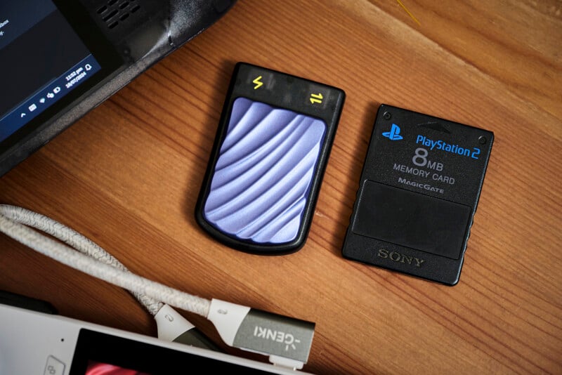 A wooden desk holds two memory cards side by side. On the left is a card with a wavy purple design and lightning bolt symbols. On the right is a black PlayStation 2 8MB memory card. Nearby are parts of a laptop and a USB hub.