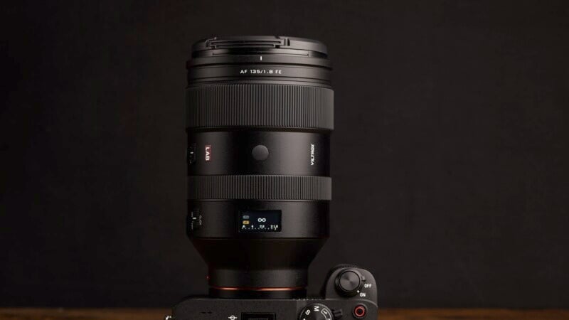 Professional camera lens mounted on a camera body against a dark background, featuring precise detailing, a focus ring, and a clear brand label. The setup is showcasing the lens's robust and high-quality design.