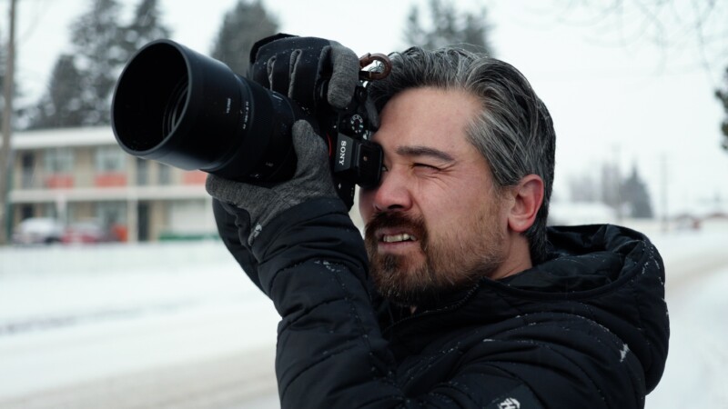 A person wearing a black jacket and gloves is holding a camera up to their face, taking a photo. They are outside on a snowy day with trees and a building in the background.