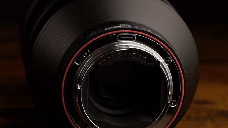 Close-up view of the rear mount of a camera lens, highlighting the metal contacts and circular opening. The outer ring has a red band, and the background is dark, emphasizing the lens details.