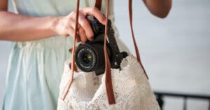 A person places a camera with a zoom lens into a white lace bag with brown straps. The person is wearing a light blue dress. Background is out of focus.