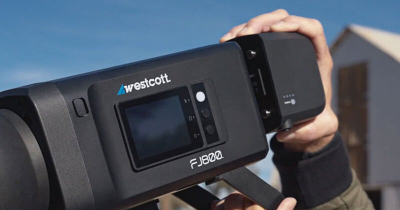 A person holds a Westcott FJ80 drone, displaying its screen and controls. The sky and part of a building are visible in the background.