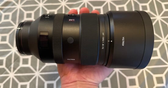 A person holding a Viltrox AF 135mm f/1.8 FE camera lens, with a geometric patterned background. The lens is large and black, featuring various dials and labels.