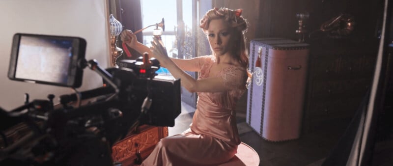 A woman in a pink dress sits on a stool, adjusting her hair in front of a camera. The lighting equipment around suggests a photo or film shoot. The room has vintage decor, including a suitcase and wooden furniture.