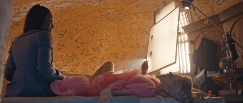 A person in a robe reclines on a bed beneath an ornate, decorated ceiling. Another person sits nearby. The room is warmly lit, with light streaming through a window, and there are various objects and books on a shelf in the background.