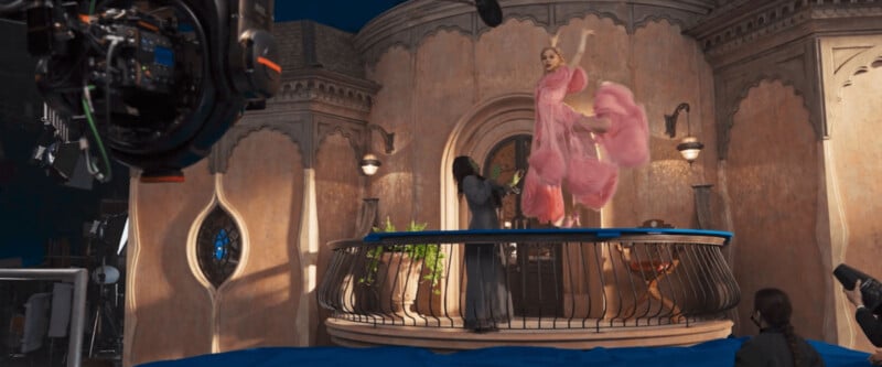 A film set with a camera capturing a woman in a flowing pink dress performing on a balcony. The backdrop includes an ornate building with arched windows, and crew equipment is visible around the scene.