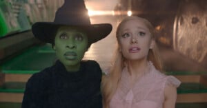 Two characters from "Wicked" stand side by side, looking upward with a surprised expression. One is dressed as the green-skinned Wicked Witch in dark attire and a hat, while the other is Glinda in a pink dress with blonde hair. Light shines behind them.