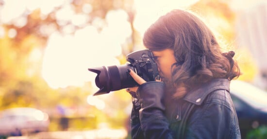 A person with long, wavy hair, wearing a black leather jacket, is holding a DSLR camera up to their eye, taking a photo outdoors during the daytime. The background is softly blurred with sunlight filtering through, creating a warm, golden glow.