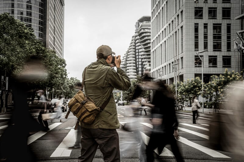 A person stands on a bustling city crosswalk, wearing a green jacket and cap, and holding a camera. The background features tall buildings and blurred pedestrians in motion, conveying a sense of urban activity.