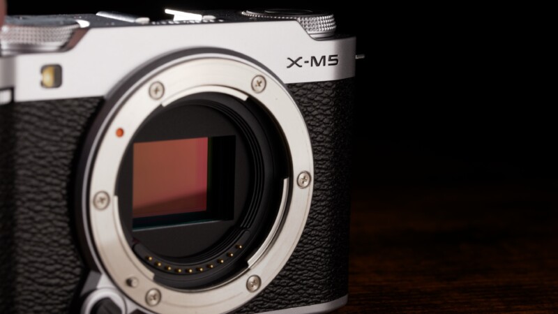 A close-up view of a silver and black digital camera with the lens removed, revealing the sensor. The camera has textured grips and the label "X-M5" near the top. The background is dark.