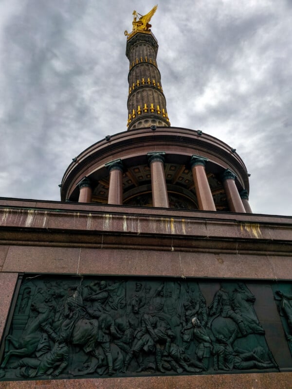 Low-angle shot of the Berlin Victory Column against a cloudy sky. The column is adorned with a golden statue on top, and features detailed reliefs depicting historical scenes near the base.