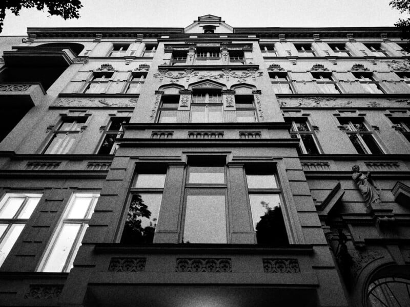 Black and white photo of a tall, ornate building featuring intricate architectural details. The perspective is from ground level looking up, showcasing large windows, decorative moldings, and balconies. The sky is visible at the top.