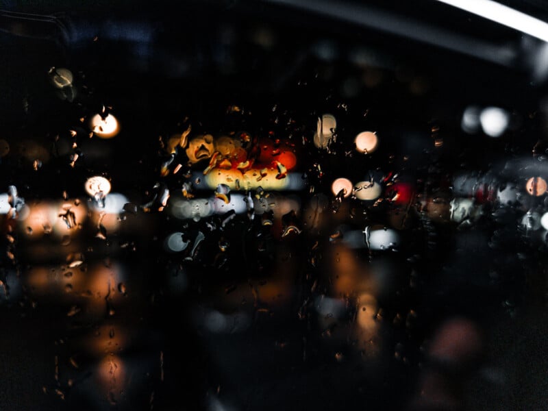 Raindrops on a window create a bokeh effect with blurry, colorful lights in the background, evoking an abstract, urban night scene.